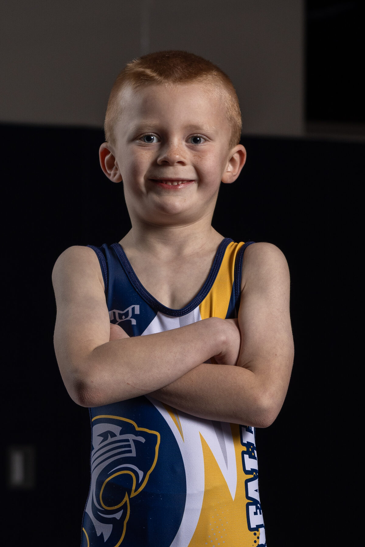 A young River Falls wrestler in a Wildcats singlet, arms folded, in the high school wrestling room